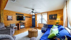 Una habitación acogedora de estilo cabaña con un sofá azul, sillones grises, una mesa de centro de madera, ventilador de techo, televisor y chimenea.
