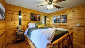 Un dormitorio de cabaña de madera con cama, mesita de noche, lámpara, ventilador de techo y cuadros en las paredes.