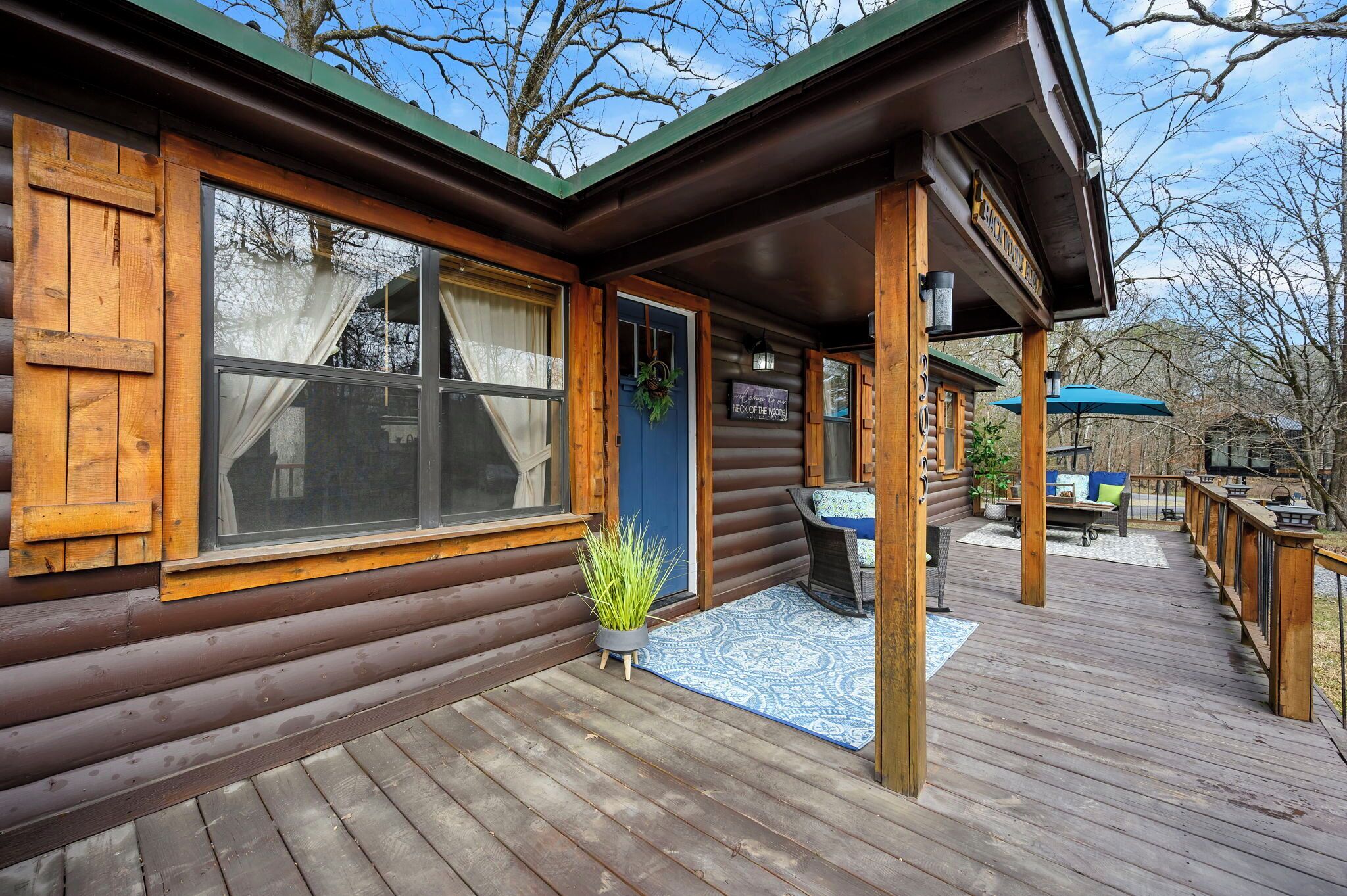 A log cabin with a porch, a sign reading "Back of the Woods," and a small table with chairs.