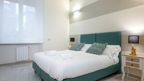 Een netjes opgemaakt bed met witte lakens en twee kussens, een nachtkastje met een lamp, en uitzicht buiten door een raam.