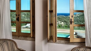 Una habitación con dos sillones a rayas, una pequeña mesa redonda y vistas a una piscina y paisaje a través de ventanas abiertas.