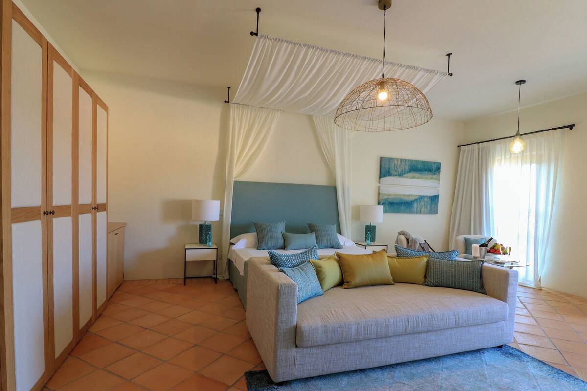 A bedroom with a bed, a sofa, a nightstand, a lamp, a hanging light fixture, and a window with curtains.