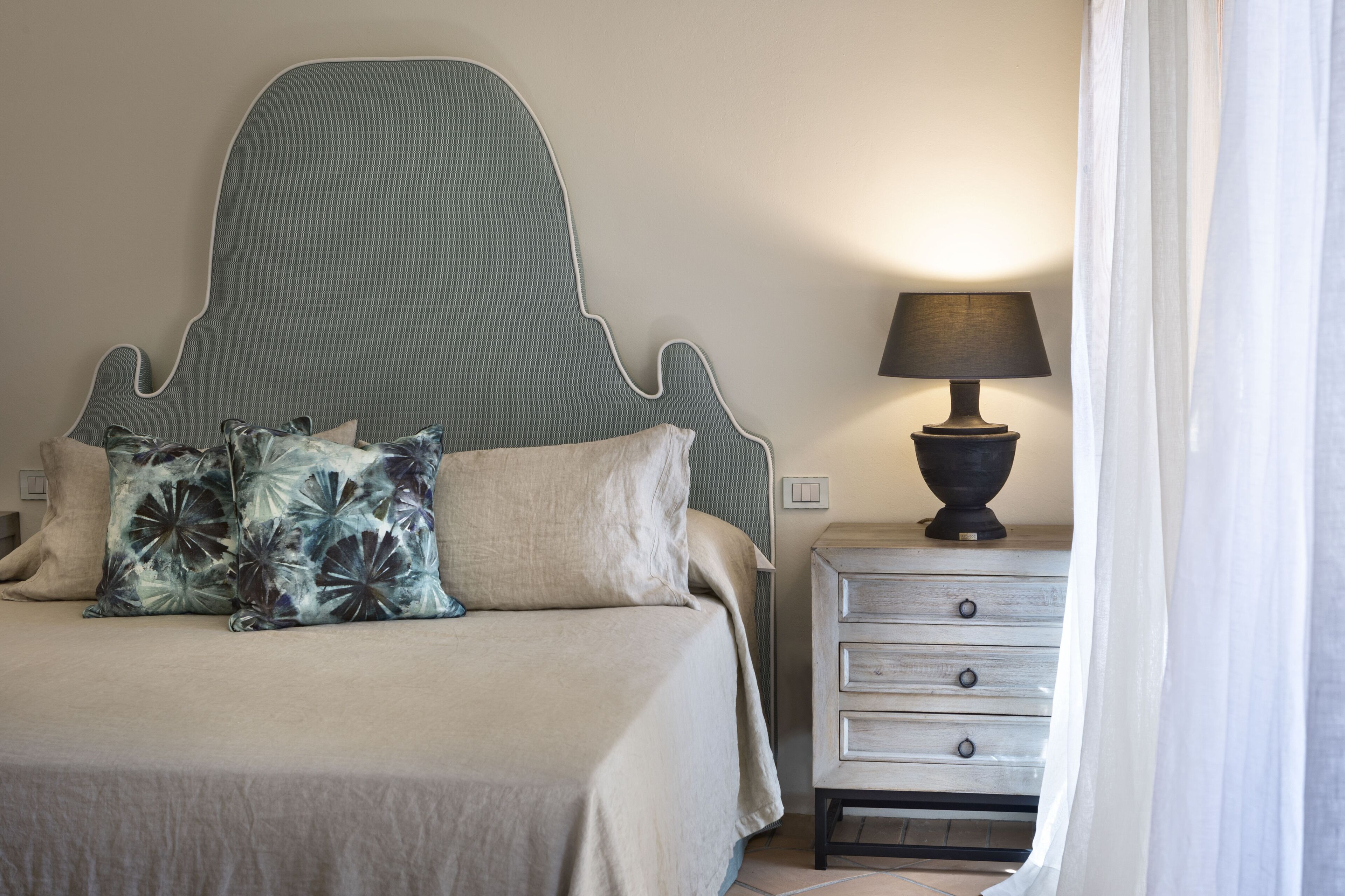 A neatly made bed with a headboard, a bedside table with a lamp, and a curtain partially drawn.