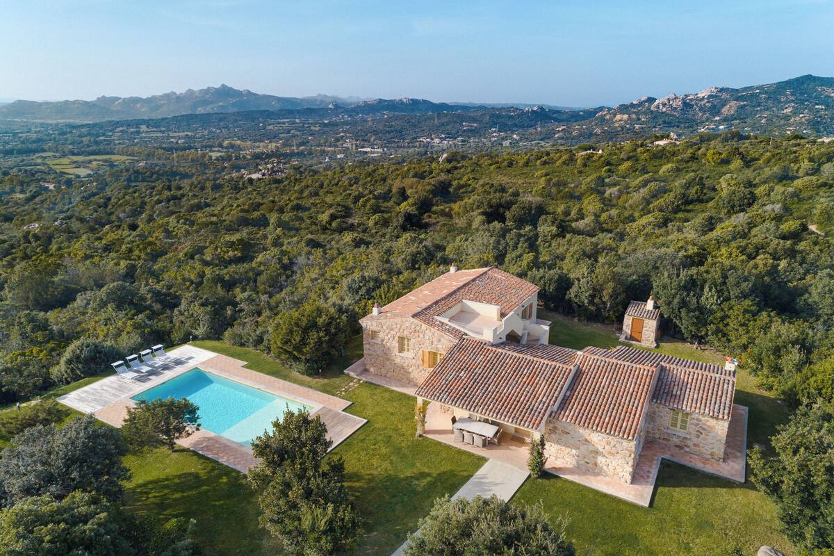 An aerial view of a stone house with a swimming pool, surrounded by lush greenery and mountains in the background.