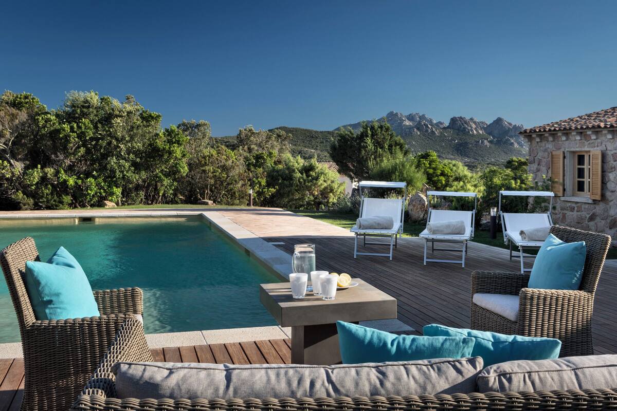 A poolside area with wicker furniture, a table with glasses and lemons, and a view of mountains.