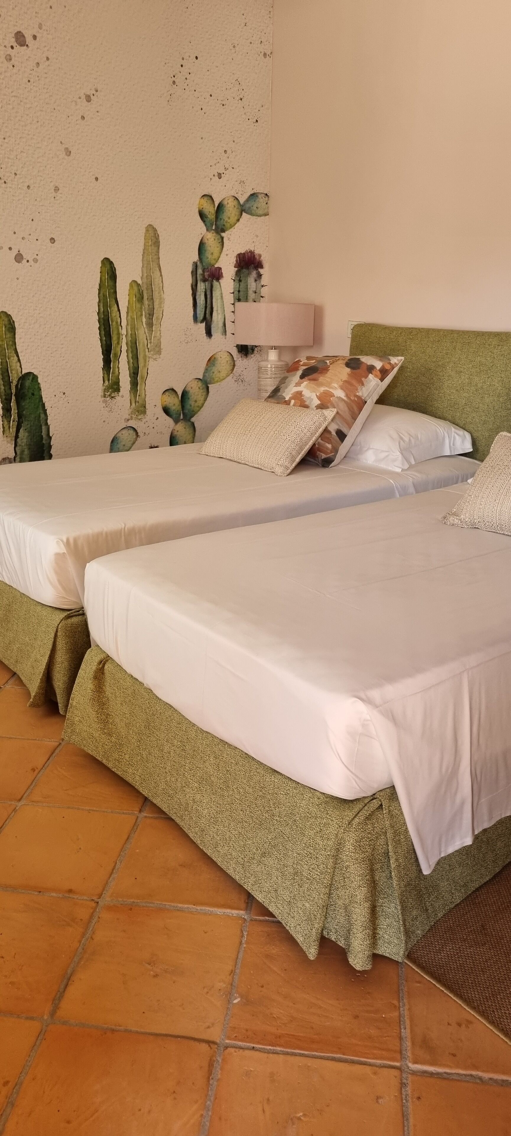 Una habitación con dos camas, cada una con ropa blanca y falda de cama verde. Hay un mural en la pared con cactus y flores, y una lámpara en la mesita de noche.