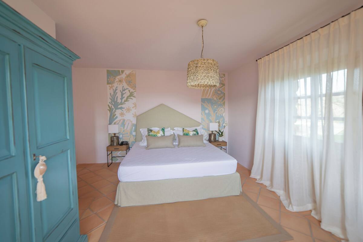 A bedroom with a bed, a nightstand, a lamp, a hanging light fixture, and a window with curtains.