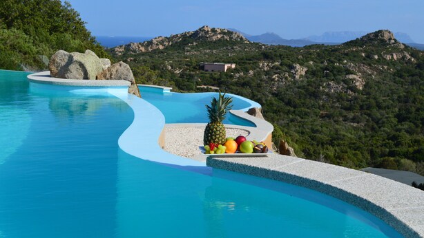 Una piscina infinita curvada con arreglos de frutas en el borde, rodeada por un paisaje natural y montañas.