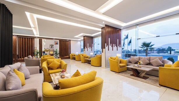 A modern hotel lobby with yellow and grey sofas, a wooden coffee table, and large windows offering a city view. - Al Qasr Hotel Suites - Al Wadiin (Ahad Rafidah)