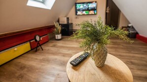 A cozy room with a wooden floor, a small round table with a remote control and a potted plant, a television mounted on the wall, and a window providing natural light. - Tiny maisonette in the center with wifi & parking space (Nordhorn)