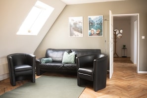 A room with a black leather sofa, two black armchairs, a green rug, and a wooden herringbone floor. There are two framed pictures on the wall, a window with a view, and a door leading to another room.