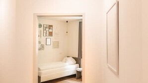 A hallway with wooden flooring leading to a bedroom with a bed and a nightstand. There is a framed picture on the wall and a potted plant by the door.