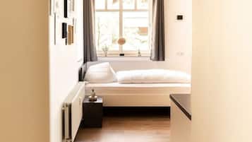 A narrow hallway leading to a bedroom with a window, a radiator, and a small bedside table.
