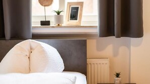 A bedroom with a bed, a window with curtains, a bedside table with a plant, and a radiator. - FAIR BED 8 City Studio with kitchen, bathroom, WLAN, & PP (Nordhorn)