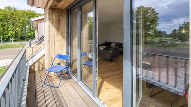 Una terraza de madera con un ventanal que da a una habitación con sofá y mesa de comedor.
