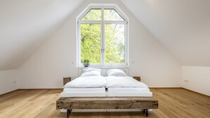 Un dormitorio con techo inclinado, un ventanal grande, estructura de cama de madera y colchón blanco.