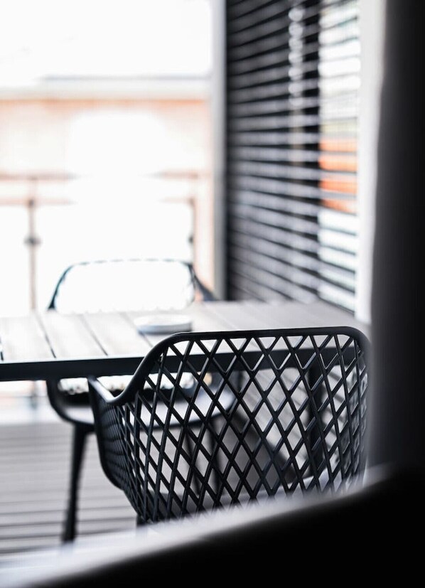 Une chaise en métal avec un dossier orné d’un motif losangique, une table en bois et une fenêtre avec des stores horizontaux.