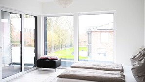 A modern bedroom with a large sliding glass door leading to a balcony, a bed with two pillows, and a small black ottoman with a cushion.
