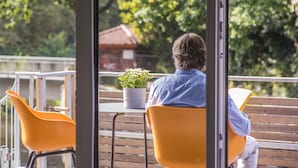A person sitting on a yellow chair at an outdoor table with a potted plant, overlooking a balcony with a view of trees and a building. - "Vechte-Garten" new build with a view of the water & PP (Nordhorn)