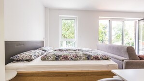 A bedroom with a bed, a grey sofa, and a wooden table. There are two windows providing natural light. - Vechte view studio, new building with balcony, WLAN & PP (Nordhorn)