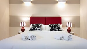Un lit bien fait, avec du linge de lit blanc et des serviettes pliées, deux lampes de chevet et une tête de lit rouge.
