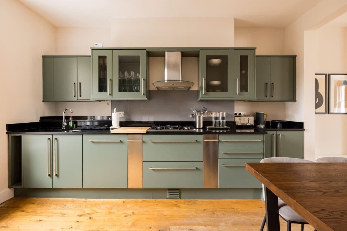 Une cuisine avec des meubles de couleur verte, une hotte de cuisinière en acier inoxydable et une table à manger en bois.