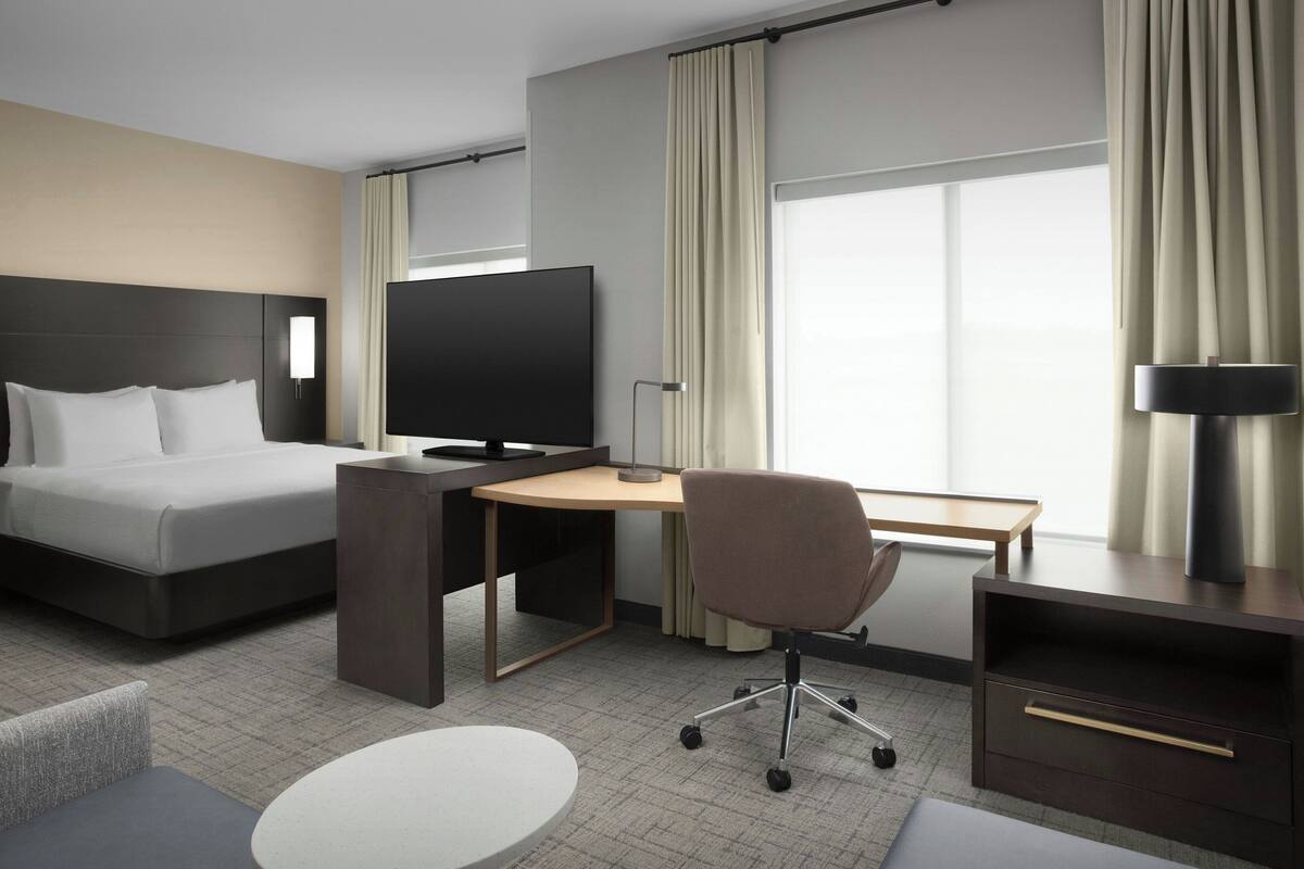 Quarto de hotel com cama, escrivaninha, cadeira, TV e janelas amplas com cortinas.
