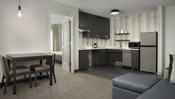 A modern kitchen with dark cabinets, a microwave, and a refrigerator. There is a dining table with chairs and a gray sofa in the living area.