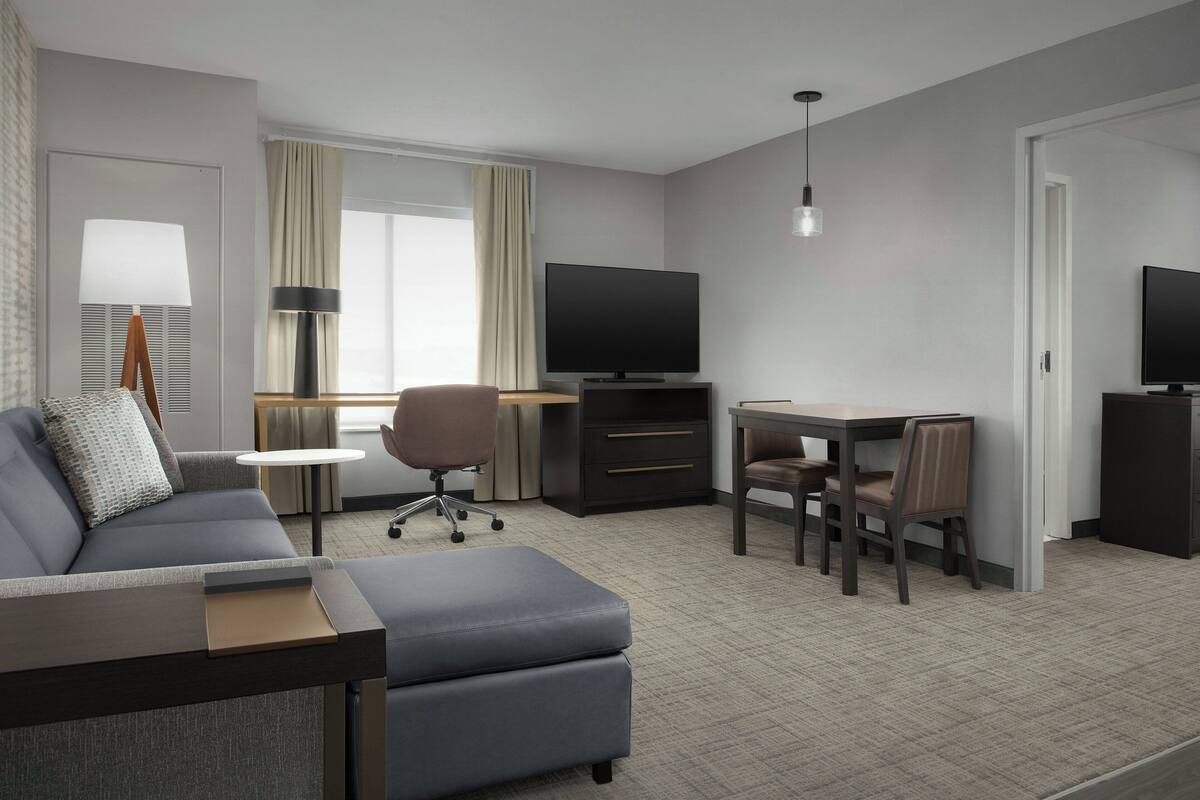 Quarto de hotel moderno com sofá cinzento, pequena mesa de jantar, televisão e escrivaninha.