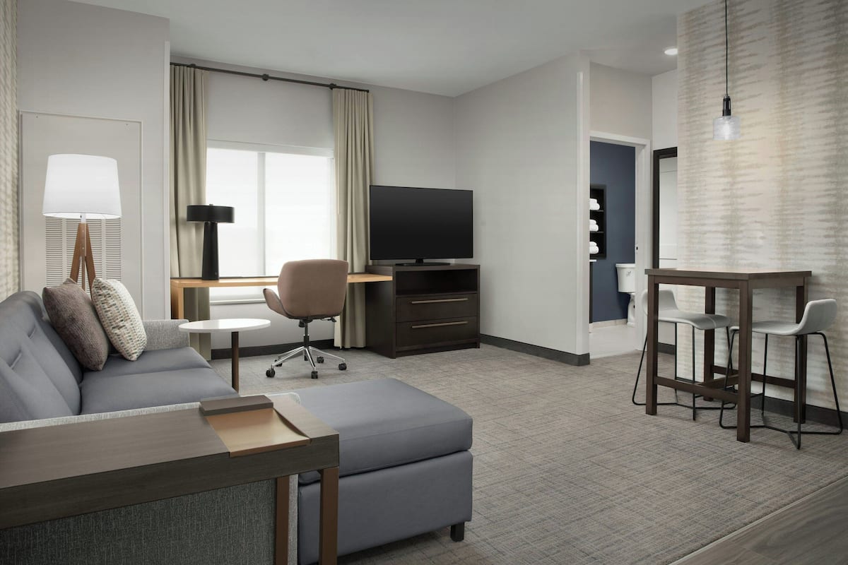 Quarto de hotel moderno com sofá, pequena mesa de jantar, televisão e área de bar.