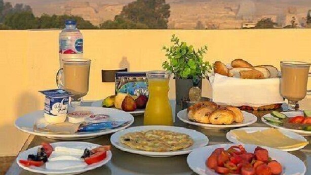 A breakfast table with various food items and drinks, set against a backdrop of pyramids. - Pyramids Heart Hotel (Giza)