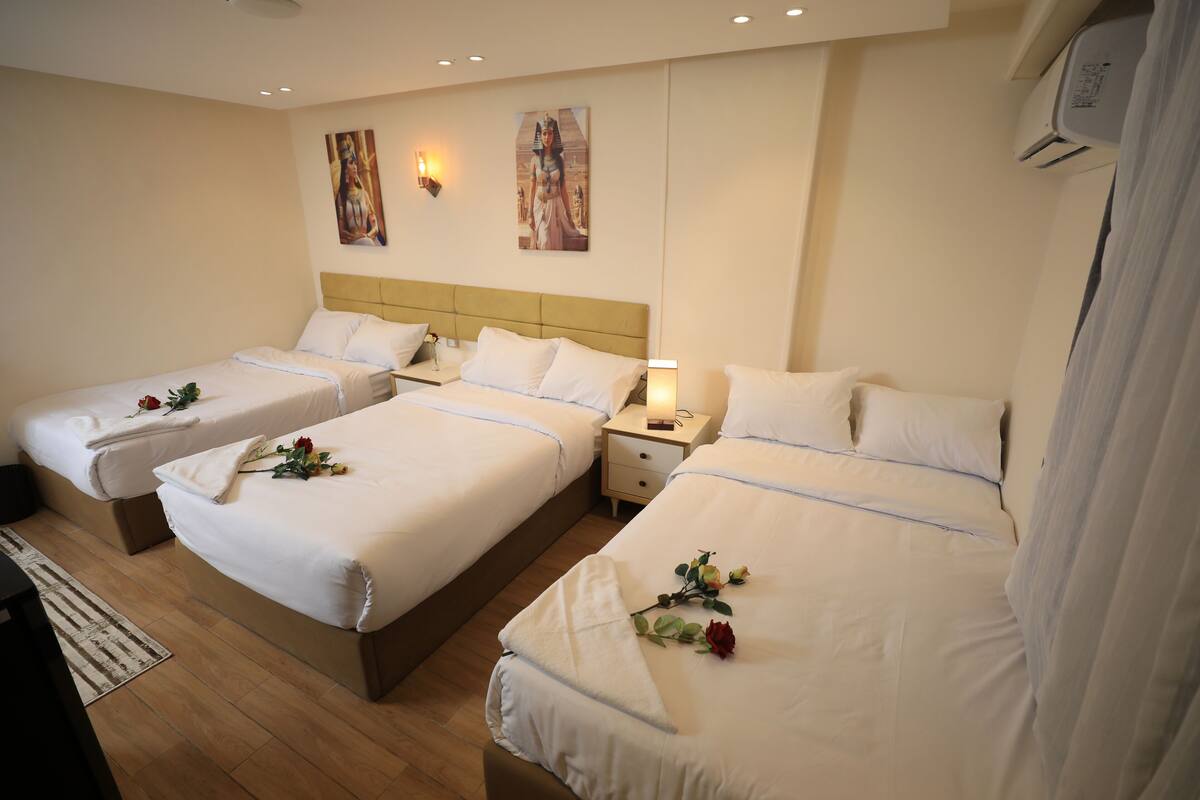 A hotel room with three beds, each with white linens and a single rose. There are wall-mounted lights and a ceiling fan.