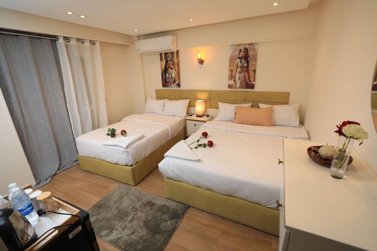 A hotel room with two beds, a desk, a chair, a small table with a vase of flowers, and a window with curtains.
