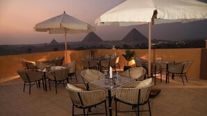 An outdoor dining area with wicker chairs and tables, white umbrellas, and a view of pyramids at dusk. - Pyramids Heart Hotel (Giza)