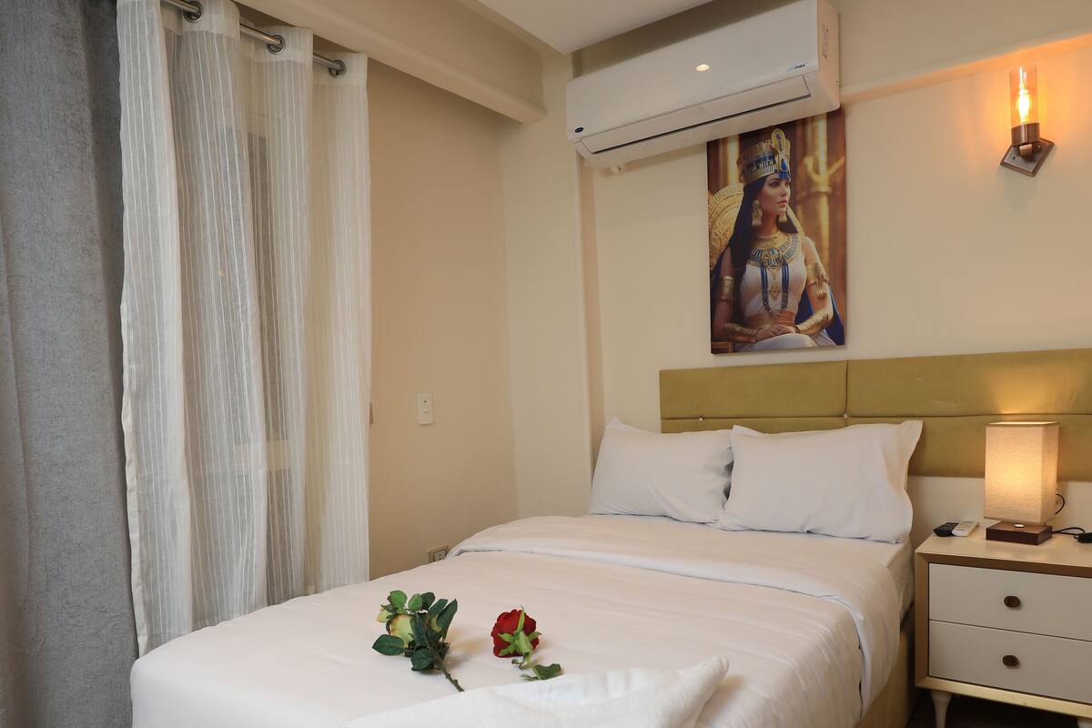 A neatly made bed with white linens, a bedside table with a lamp, and an air conditioning unit mounted on the wall.
