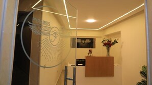 A reception area with a glass wall displaying the logo 'PYRAMIDS' and a logo with an eye and wings. There is a wooden reception desk, a vase with flowers, and a framed picture on the wall. - Pyramids Heart Hotel (Giza)