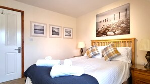Ein ordentlich bezogenes Bett mit weißen und karierten Kissen, ein hölzernes Kopfteil und ein Nachttisch mit einer Lampe. Darüber an der Wand hängen drei gerahmte Bilder.