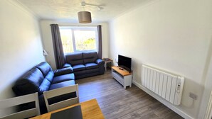 A compact living space with a sofa, a TV on a wooden stand, a radiator, and a window with curtains. - Beach front apartment with direct beach access! (Perranporth)