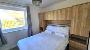 A bedroom with a wooden headboard, a large window with curtains, and a neatly made bed. - Beach front apartment with direct beach access! (Perranporth)