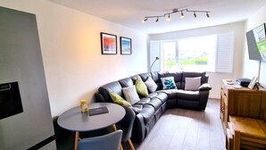 A modern living room with a sectional sofa, a small round table, and a flat-screen TV mounted on the wall. - Holiday apartment with direct access to Perranporth beach! (Perranporth)