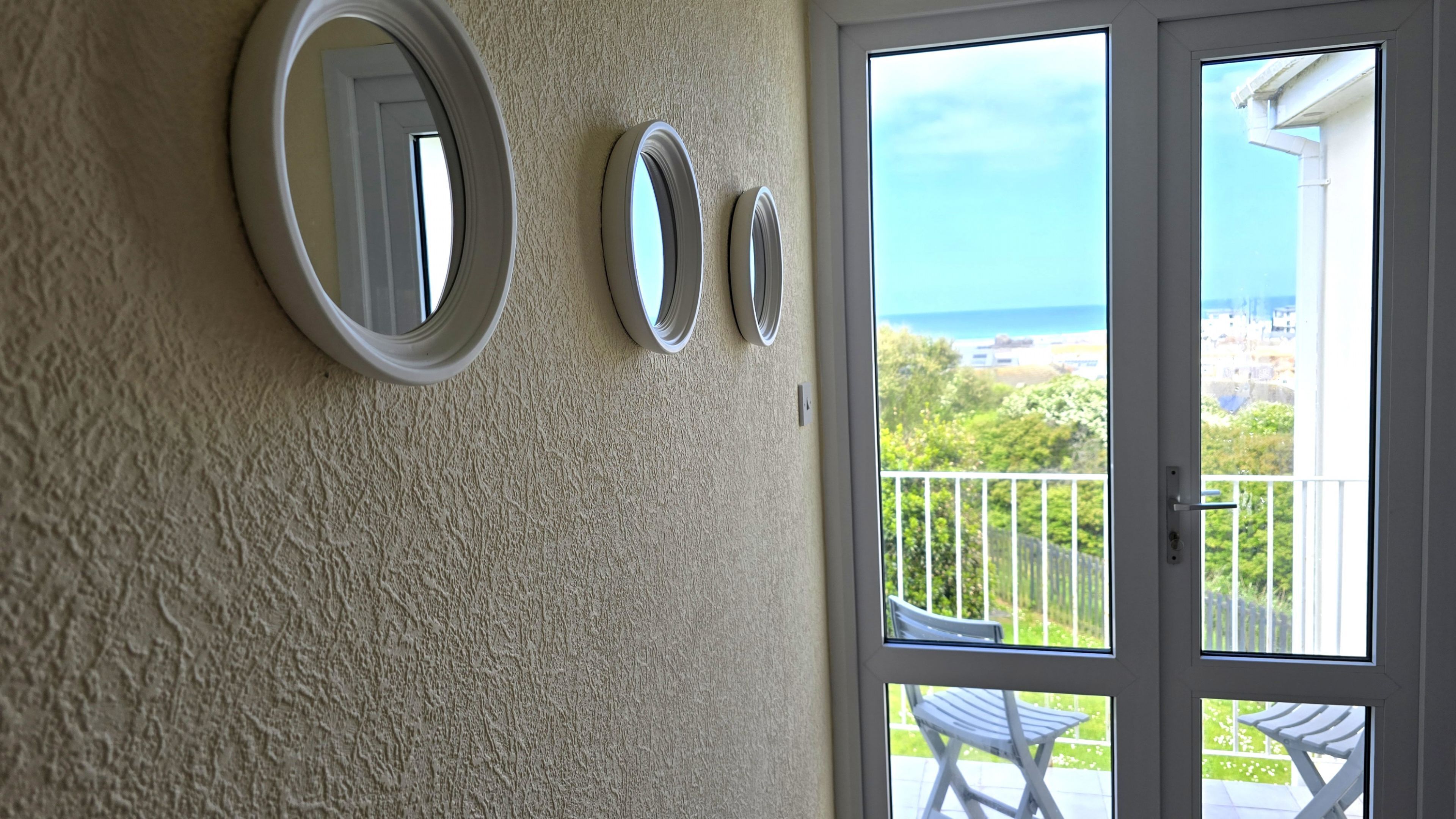 A room with three wall-mounted mirrors and a sliding glass door leading to a balcony with a view of the sea.