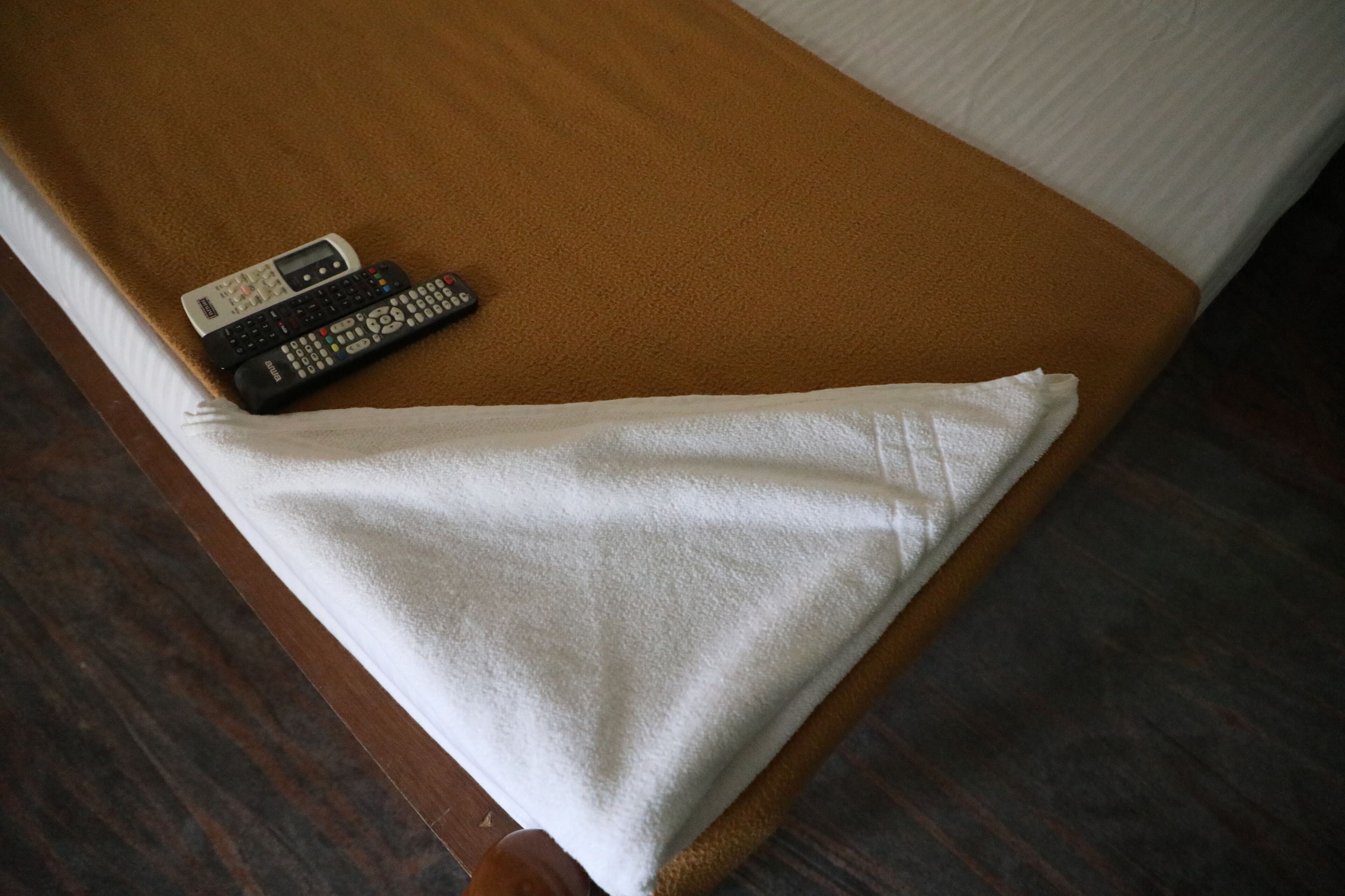 Una cama con cabecera marrón, almohada blanca y dos controles remotos sobre la cama.