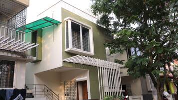 Un moderno edificio residencial de dos pisos con un balcón, una entrada protegida por un muro y un árbol en primer plano.