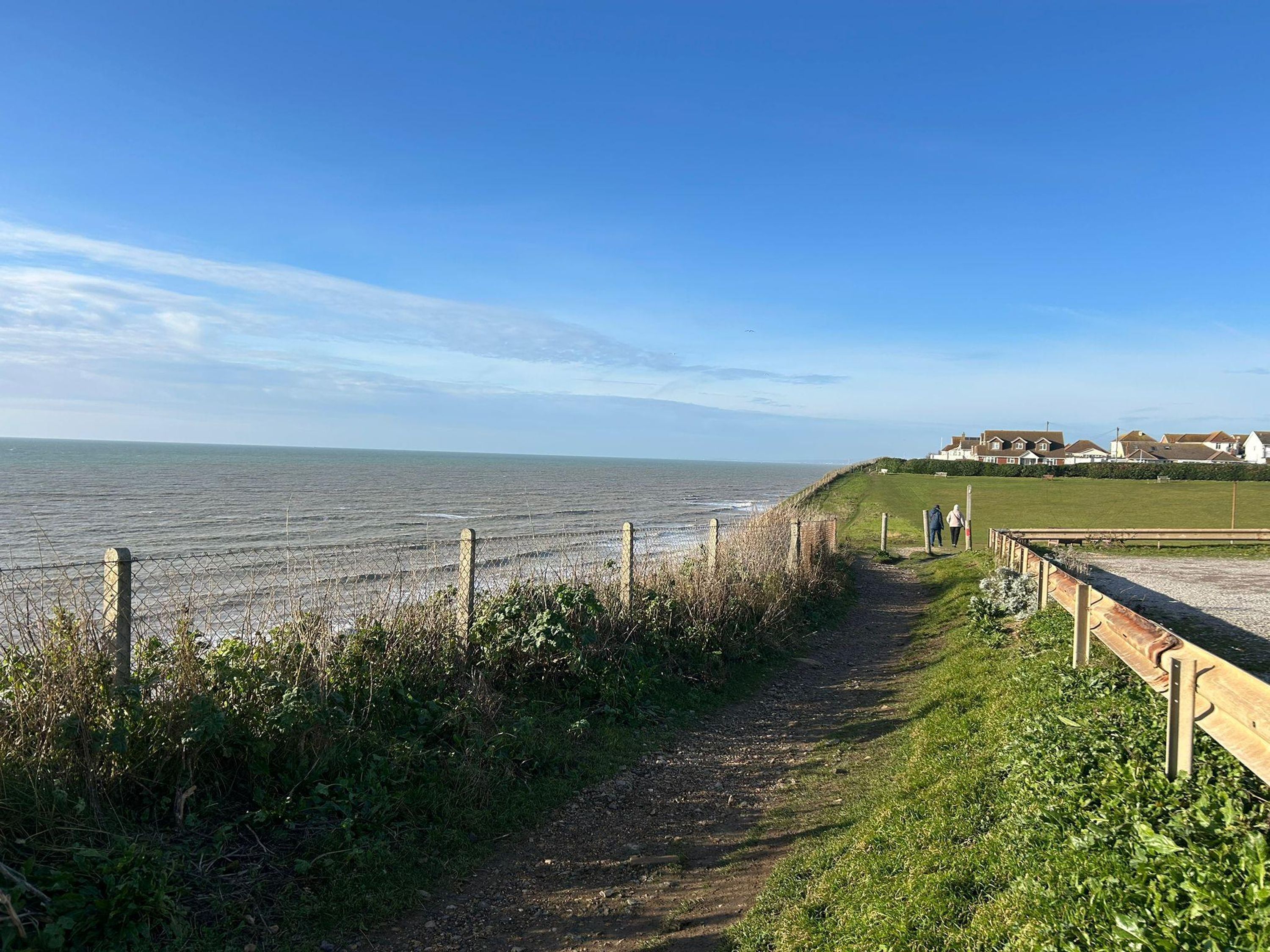 A coastal path with a fence, grassy areas, and houses in the background.