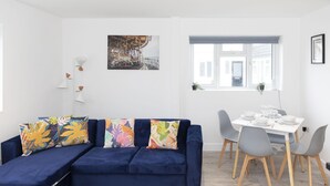 A modern living room with a blue sofa, colorful cushions, a glass coffee table, and a dining area with a white table and grey chairs. - Ocean Side With Free Parking By My Getaways (Peacehaven)