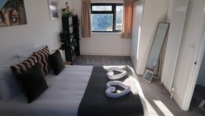 A neatly made bed with a black bedspread and white heart-shaped pillows, a large window with curtains, a plant, and a mirror on the wall. - Lovable Lancing With Garden Parking (Lancing)