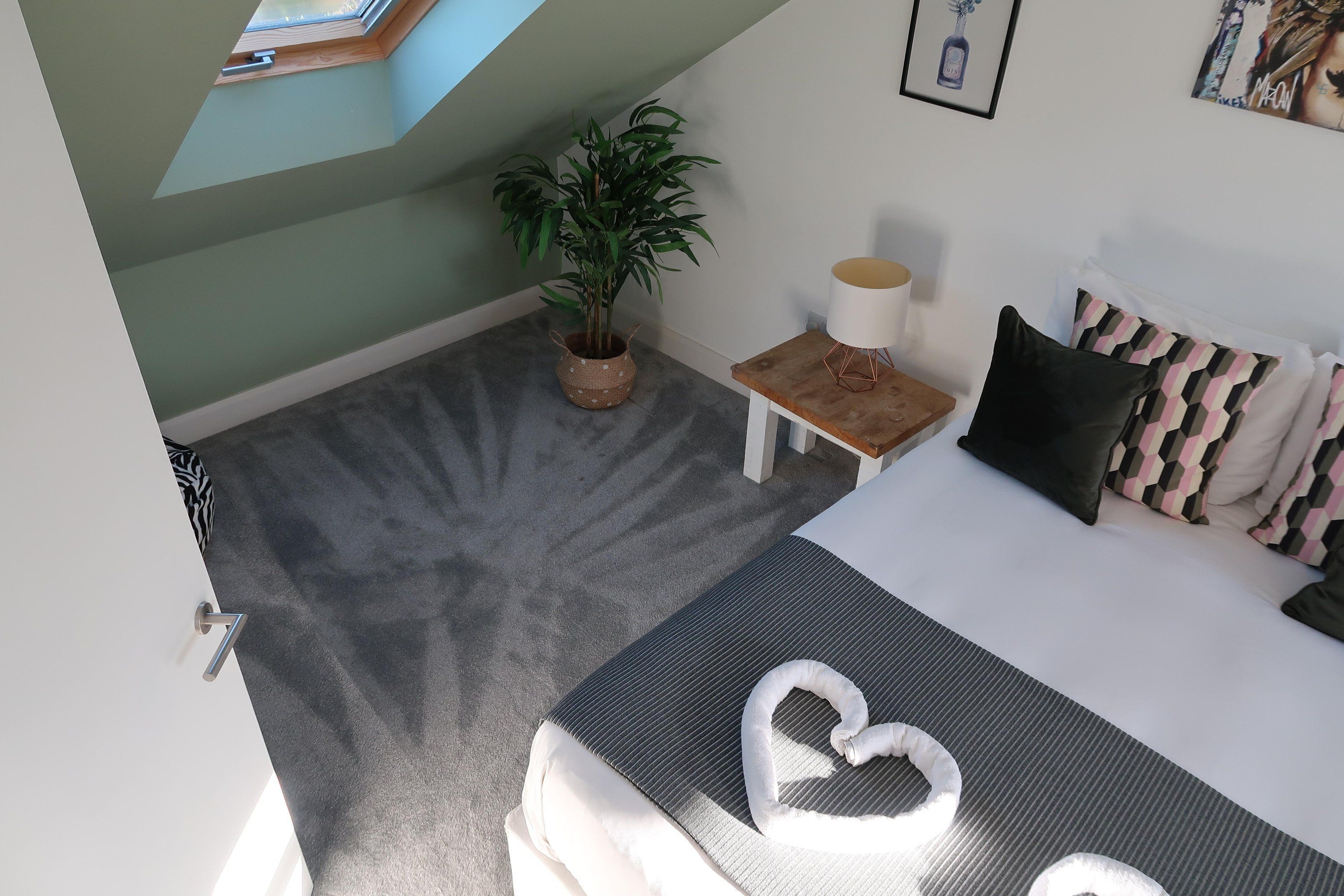 A room with a bed, a small table with a lamp, a potted plant, and a view of the sky through a skylight.