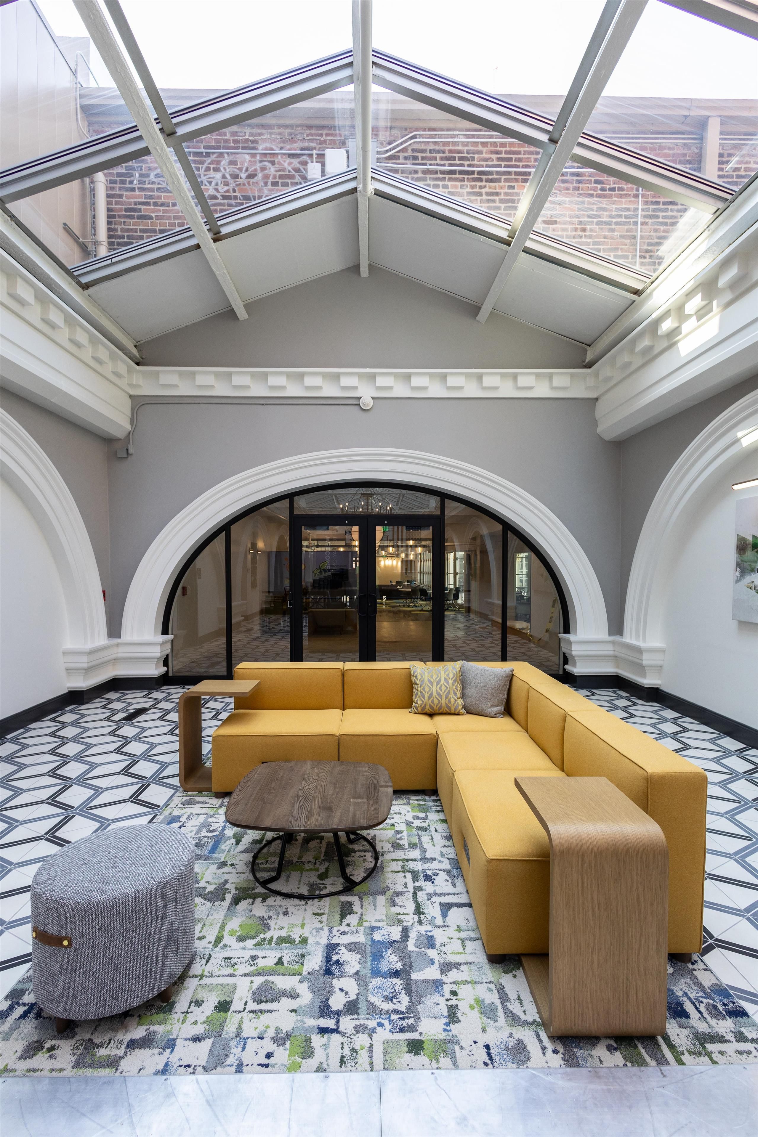 A spacious lobby with a yellow sectional sofa, a round wooden coffee table, and a grey ottoman. The floor has a patterned tile design, and the space is under a glass ceiling with a view of a brick building outside.