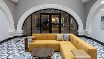 A spacious lobby with a yellow sectional sofa, a round wooden coffee table, and a grey ottoman. The floor has a patterned tile design, and the space is under a glass ceiling with a view of a brick building outside.