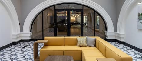 A spacious lobby with a yellow sectional sofa, a round wooden coffee table, and a grey ottoman. The floor has a patterned tile design, and the space is under a glass ceiling with a view of a brick building outside.